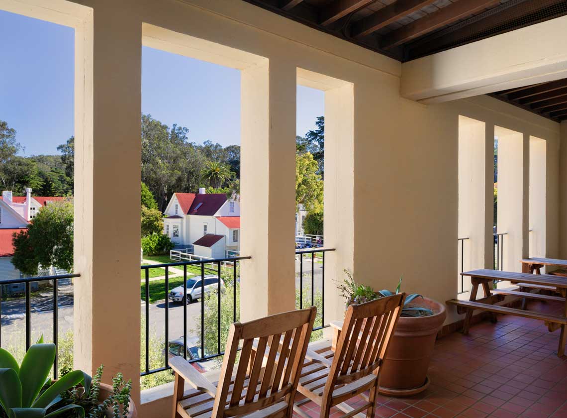 Daytime exterior view of Mesa Street from 2nd floor veranda with wooden chairs overlooking other Presidio structures