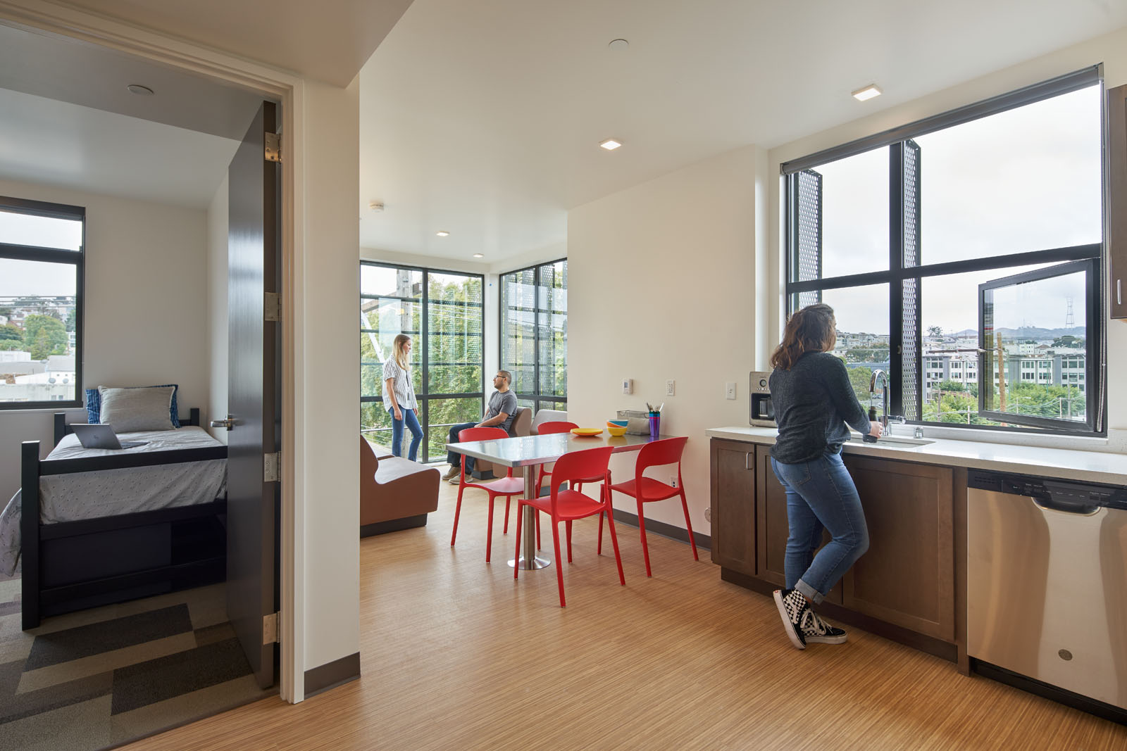 Image of student unit kitchen and dining area and entrance to bedroom. Student looking out window.