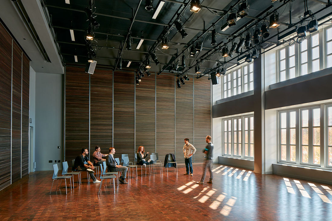 Rehearsal room with wood floors and daylight through large windows; people seated in loosely arranged chairs.