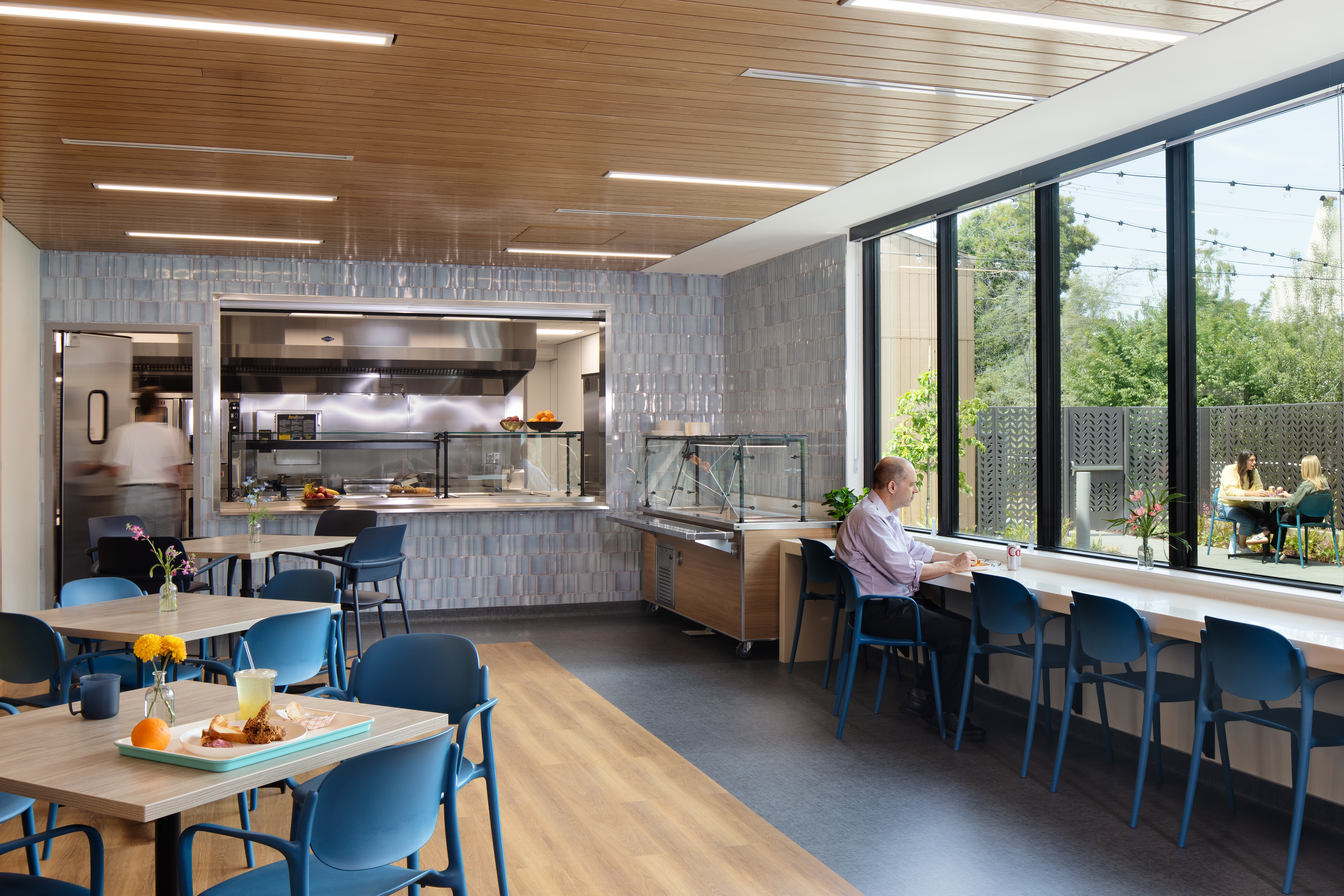 A bright cafeteria with a wood-slat ceiling and a service window into a stainless-steel kitchen, its surrounding wall covered in glossy blue-gray tile. A man eats at a long counter beside floor-to-ceiling windows overlooking a patio strung with lights, where two people sit at a table. Blue chairs and light wood tables fill the foreground, one set with a breakfast tray.