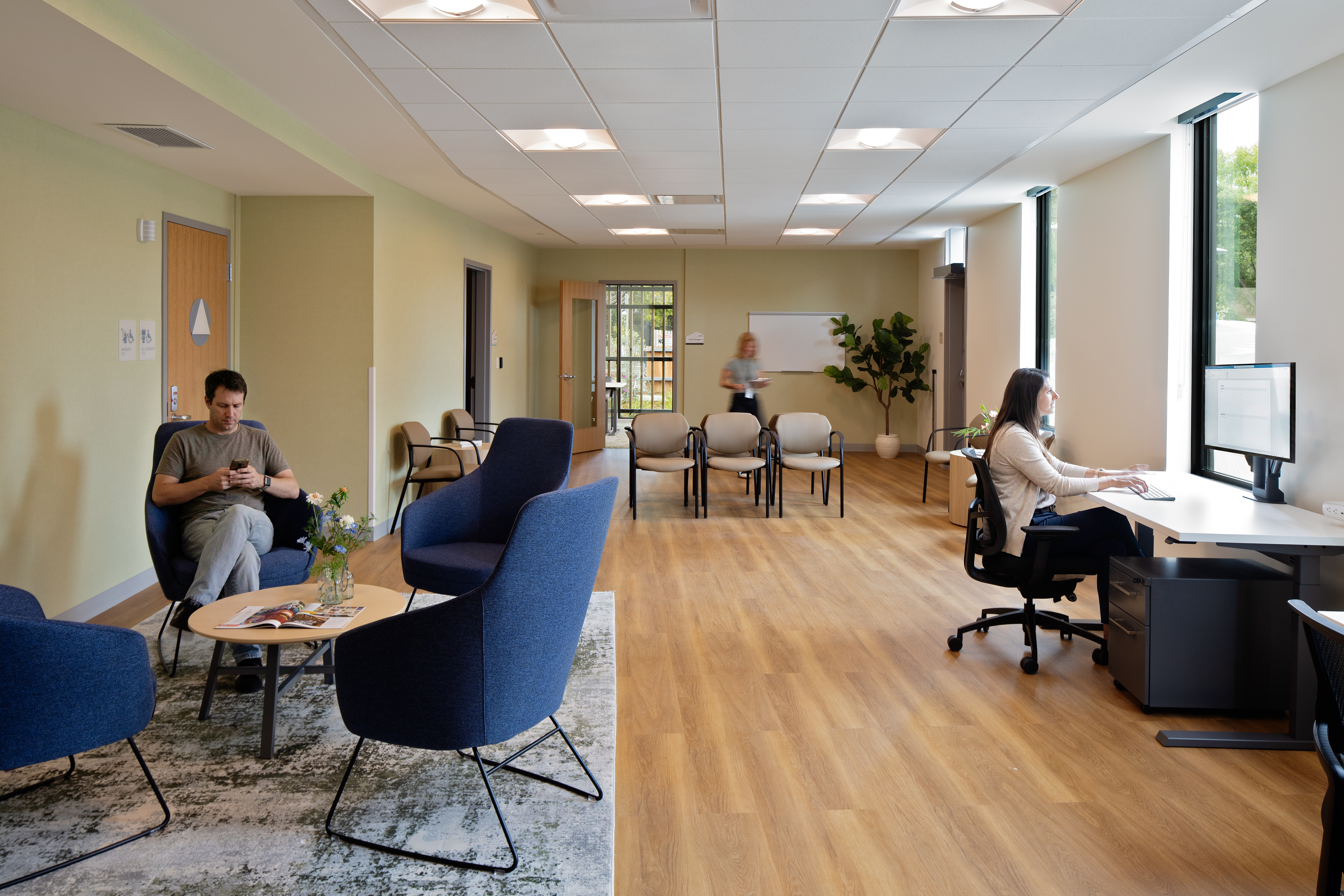 A long room with pale green walls and wood-look flooring. At left, a man checks his phone in a navy lounge chair near a low table with flowers. Rows of tan stacking chairs sit at center by a whiteboard, where a woman stands writing. At right, a woman works at a height-adjustable desk with dual monitors beside tall windows.