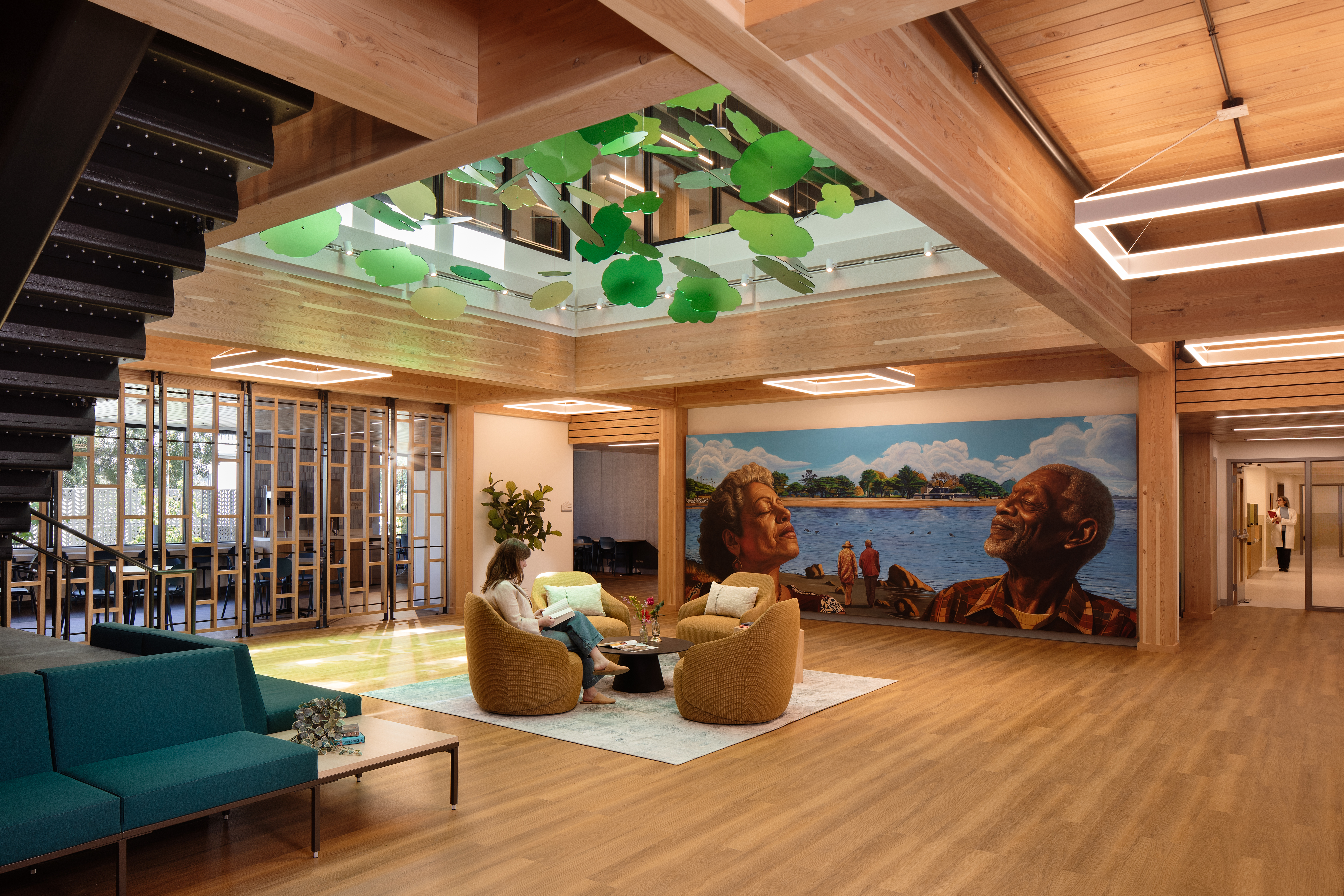 A double-height lounge with wood beams and hanging green leaf panels overhead. A woman reads in a mustard-yellow armchair by a round black coffee table on a light rug. At right, a wall mural shows an older woman and man in profile facing each other across a lake, with two figures walking the shore between them. A teal sofa sits at left; a wood-and-glass partition opens to a meeting room beyond.