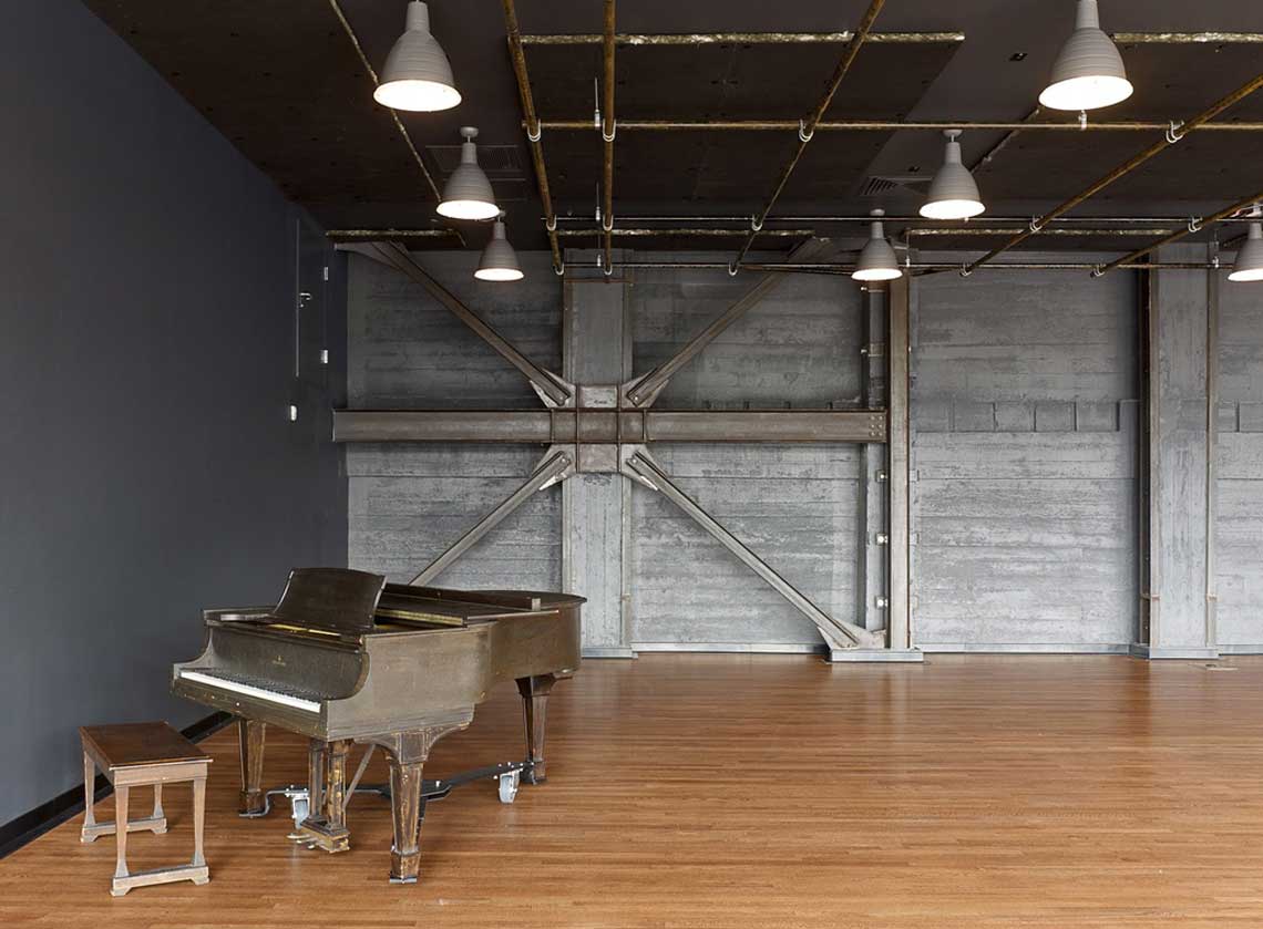 Rehearsal space with hardwood floors and exposed industrial wall and ceiling details; a piano in the foreground