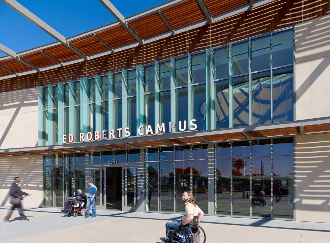 Exterior daytime view of building entrance with prominent sign on overhang; people coming and going including wheelchair users