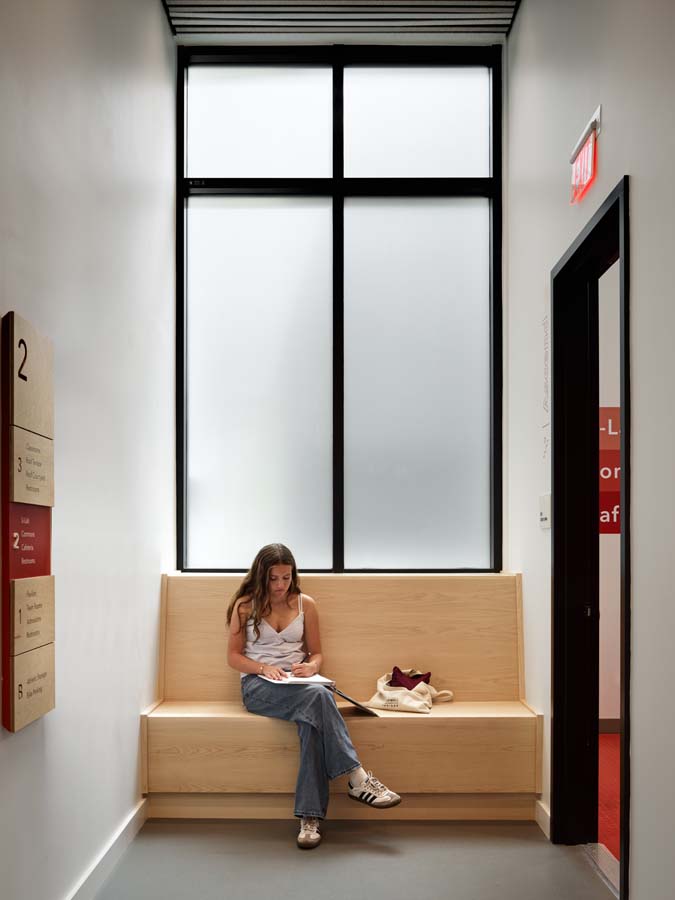 Student seated at small bench in common area hallway with frosted window behind.