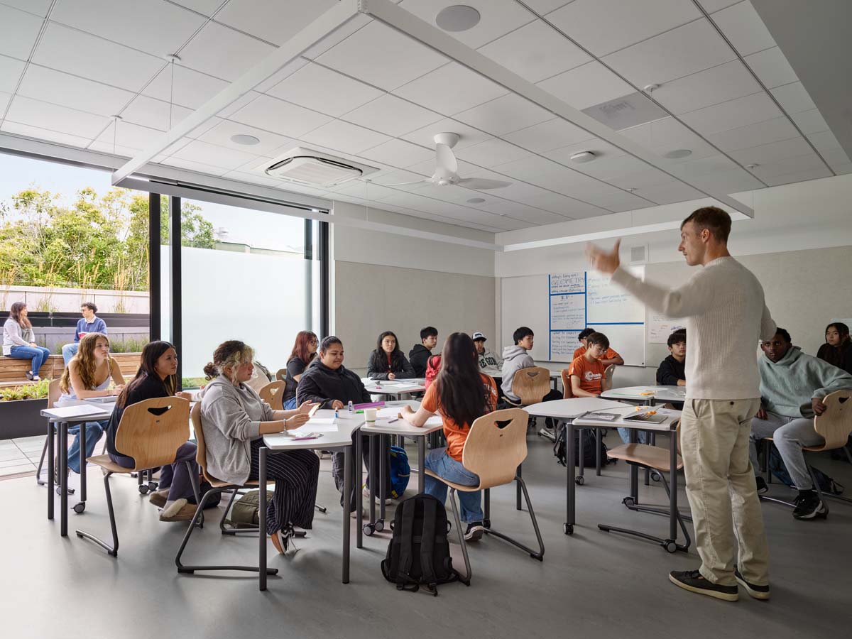 Windows behind students seated at desks in classroom with teach standing. 