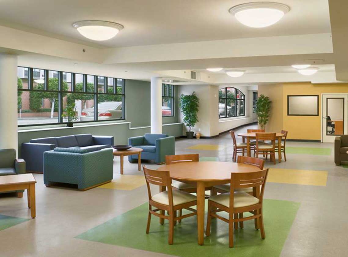 Interior community space with tables and arm chairs. Colorful green and yellow shapes accent the tile floor