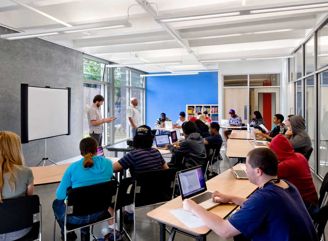 Students seated at tables with laptops in a bright classroom space with window and a blue accent wall