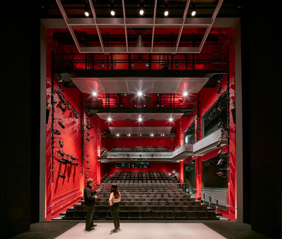 Interior view of black theater space as seen from the back of stage looking toward the house. Two people stand on stage talking.